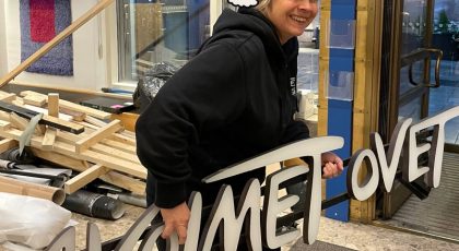 Teatterinjohtaja Hanna kantaa iloisena Teatteri Avoimien Ovien valokylttiä teatterin aulan ovesta ulos. Hänelle on piirretty päähän tonttulakki. Taustalla muuttoromua.