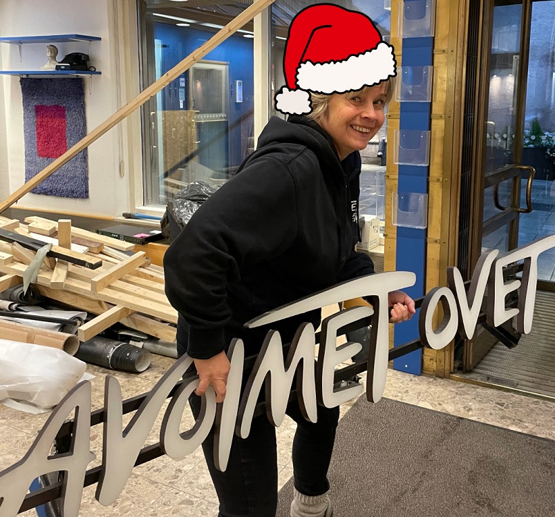 Teatterinjohtaja Hanna kantaa iloisena Teatteri Avoimien Ovien valokylttiä teatterin aulan ovesta ulos. Hänelle on piirretty päähän tonttulakki. Taustalla muuttoromua.